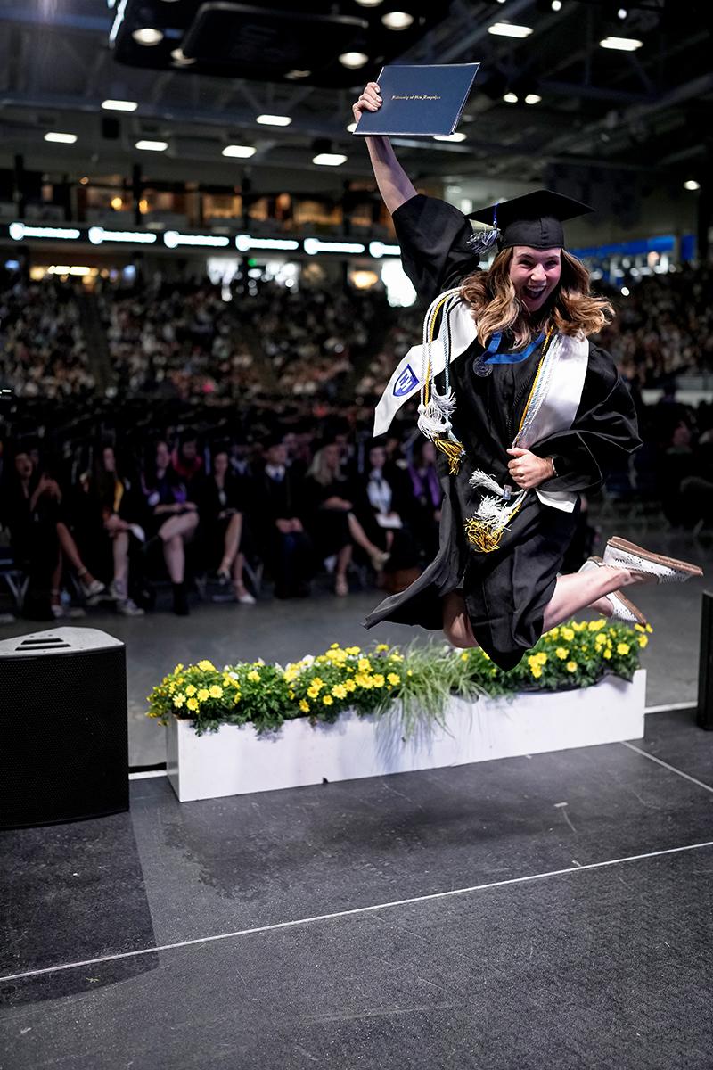 A woman graduate leaps into the air while crossing the stage at commencement
