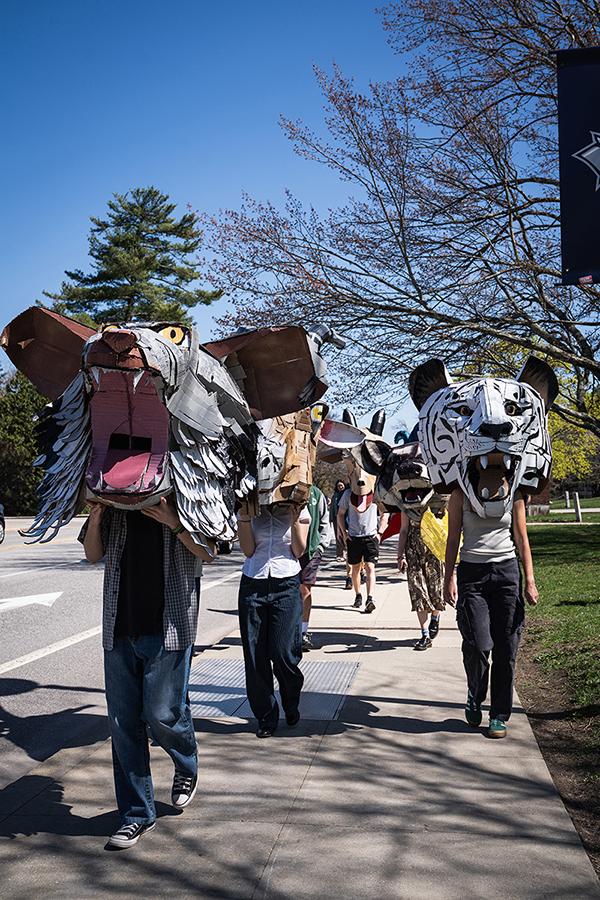 Students wear large masks created in their sculpture class during a campus parade