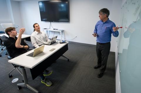 two people sitting at a table with laptops and one person standing by a whiteboard in the UNH Entrepreneurship Center