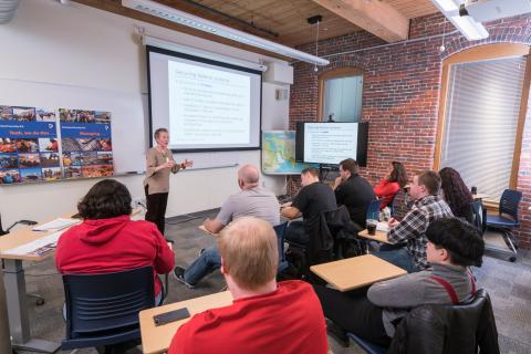 Professor standing in a classroom giving a lecture with a slide about securing federal systems in the background