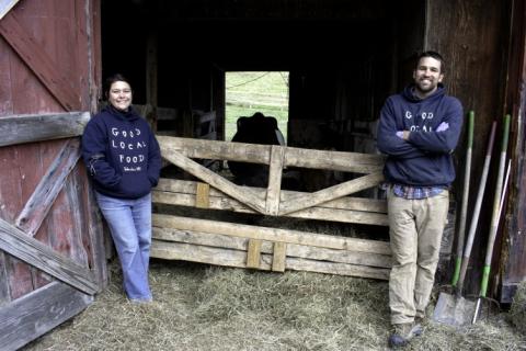 A woman and a man in dark hooded sweatshirts stand in front of the door to a barn