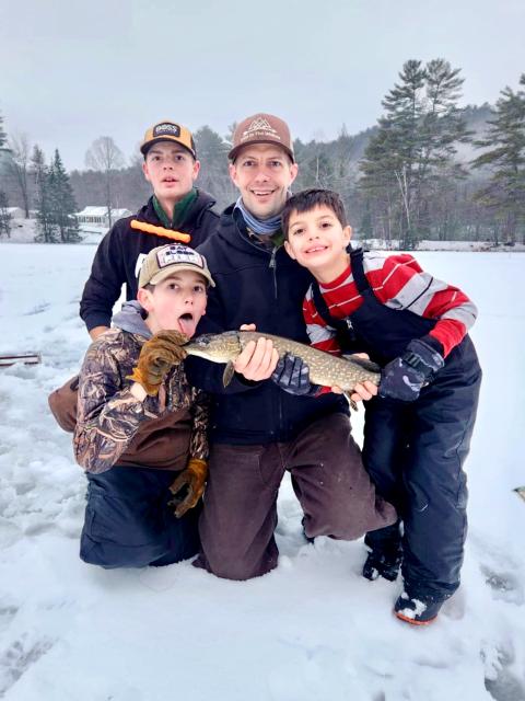 A group of three boys and one man pose with a fish atop a frozen lake