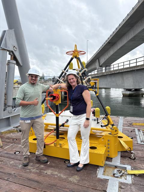 Man and woman wearing hardhats stand beside a large yellow piece of oceanographic equipment