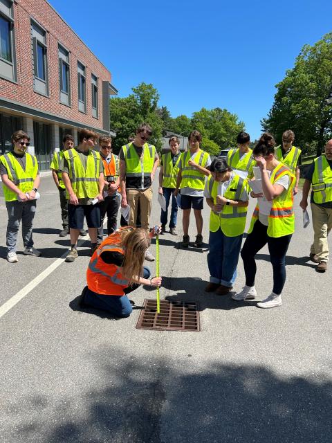 Students wearing high-viz vests stand watching an instructor measure a storm drain