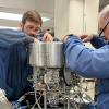 Two men in blue work shirts use tools to work on a silver metallic-looking component to be included in a rocket launch