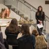 An audience is seated in front of a woman presenting alongside a series of sculptures