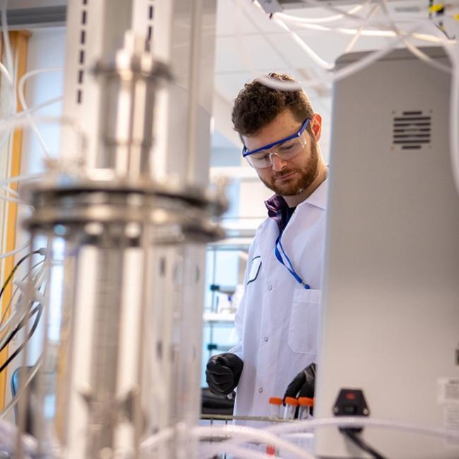 male student in lab coat standing in between lab equipment