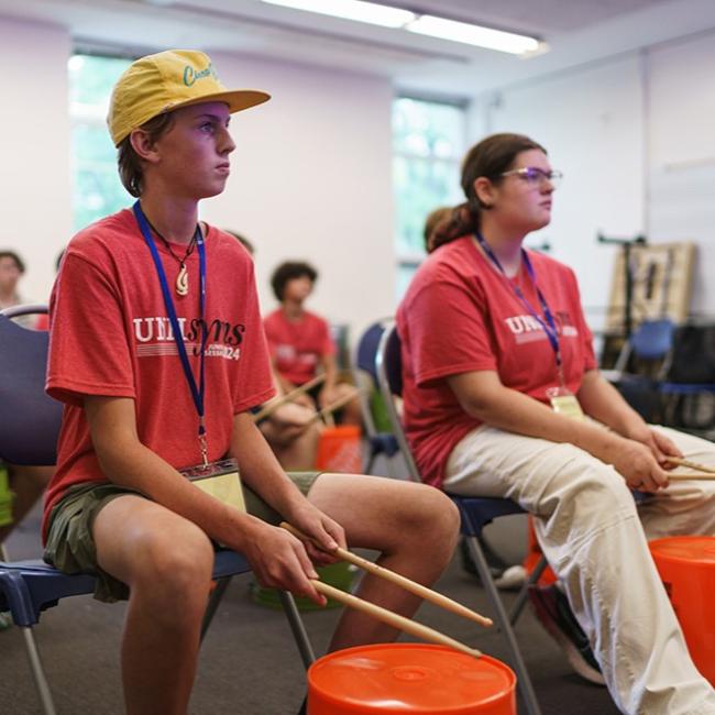 Two children sitting in front of a bucket drum holding drumsticks'