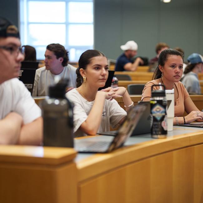Two female students sitting at a desk in a classroom with their computers sitting in front of them