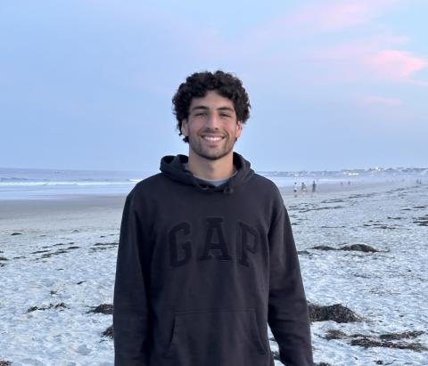 a young man with dark curly hair and a beard in front of the ocean