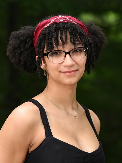 Female student with red bandanna, glasses, black tank standing in front of a backdrop of trees.