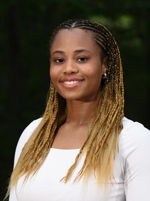 Female student in white with braided hair standing outside with trees in the background.
