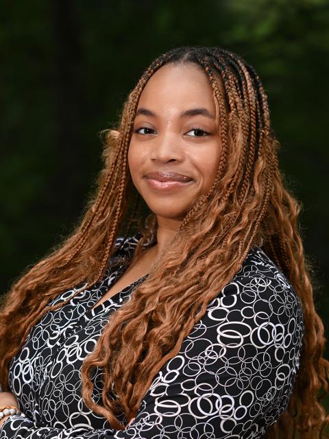 Female student with long, light brown, braided hair, black and white patterned button down shirt, standing outside with trees in the background.