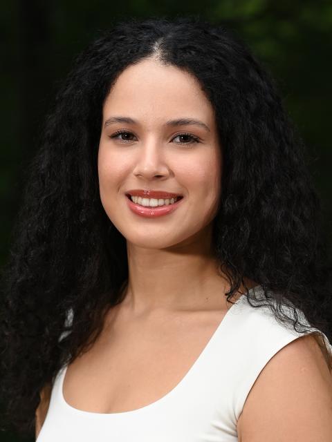 Female student with dark curly hair dressed in white standing outside with trees in the background.