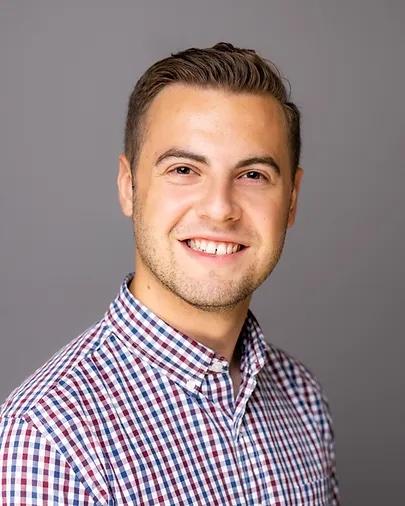 Professional headshot of George Anderson smiling wearing a red, white, and blue checkered collared shirt. 