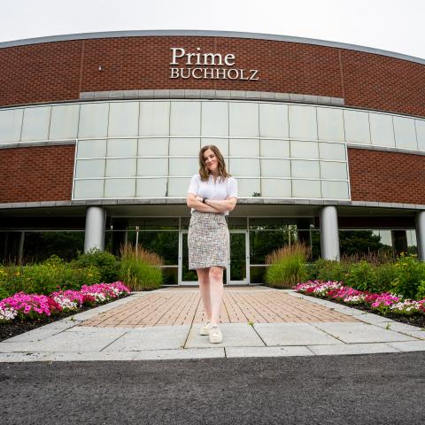 Jill standing with her arms crossed and smiling in front of the Prime Buccholz building. There are flowers aligning the walkway to the door in the background.