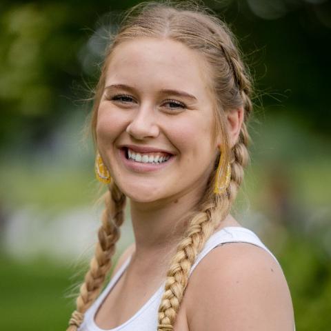 Headshot of Briella Hirsch smiling, wearing Dutch braided pigtails, white tank top, and lemon wedge ear rings.