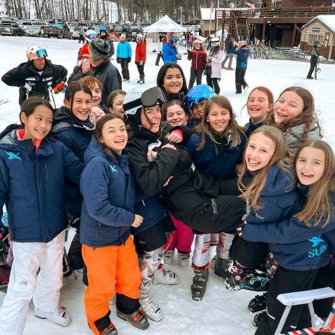 Jill Durkin wearing ski goggles, helmet, and winter clothes, being held up by a bunch of children with a ski resort and snow in the background.