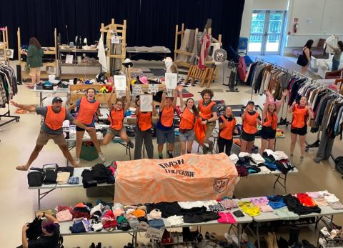 people in orange vests in a line in a large room with tables of items and clothing on racks