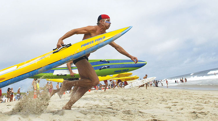 UNH alumnus Greg Johnson ’83 running with a surfboard at the beach (photo: Cape Cod Times)
