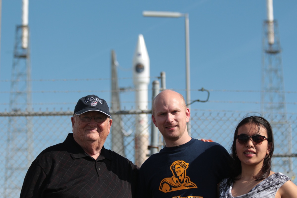 Matthew Argall stands between an elderly man and a young, dark-haired woman in front of a rocket launch for the MMS mission