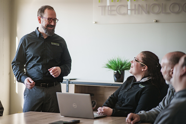 A man in glasses laughs while speaking to two colleagues in an office setting