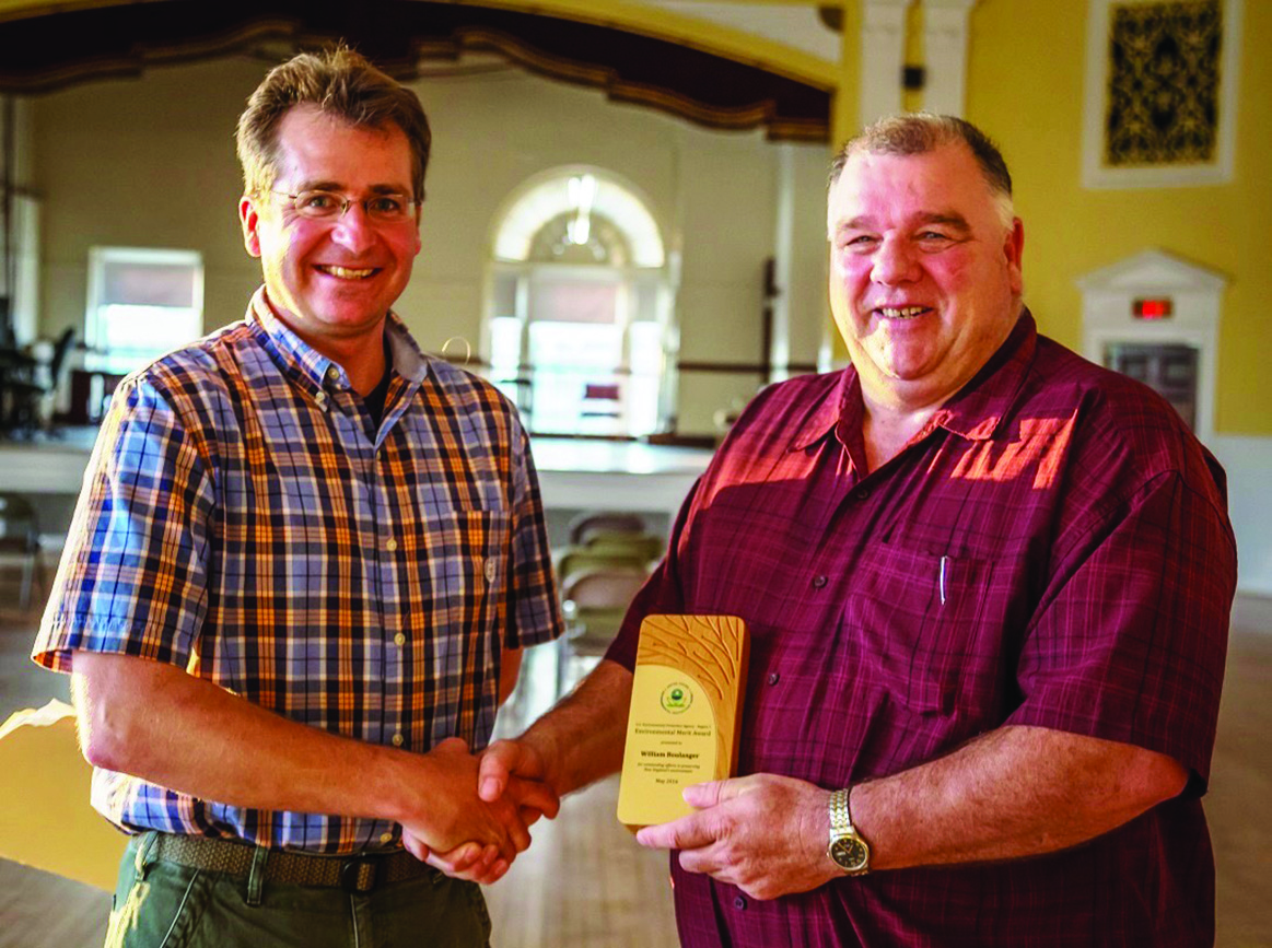 Jamie Houle and Bill Boulanger shake hands and hold a plaque