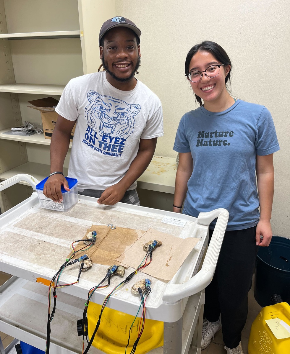 A photo of two students smiling and looking into the camera. In front of them is a workbench covered with oysters hooked up to biosensors.