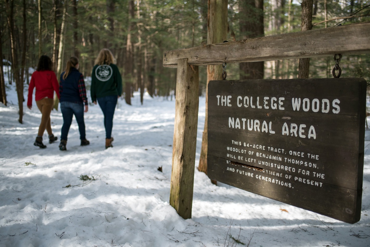 A sign in the foreground reads “The College Woods Natural Area.” Snow is on the ground and three people walk in the background through the woods.