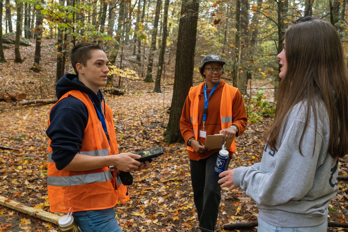Three students stand in College Woods in the fall time. Two students, pretending to survey a third student, wear bright orange vests. The third student wears a grey sweatshirt.