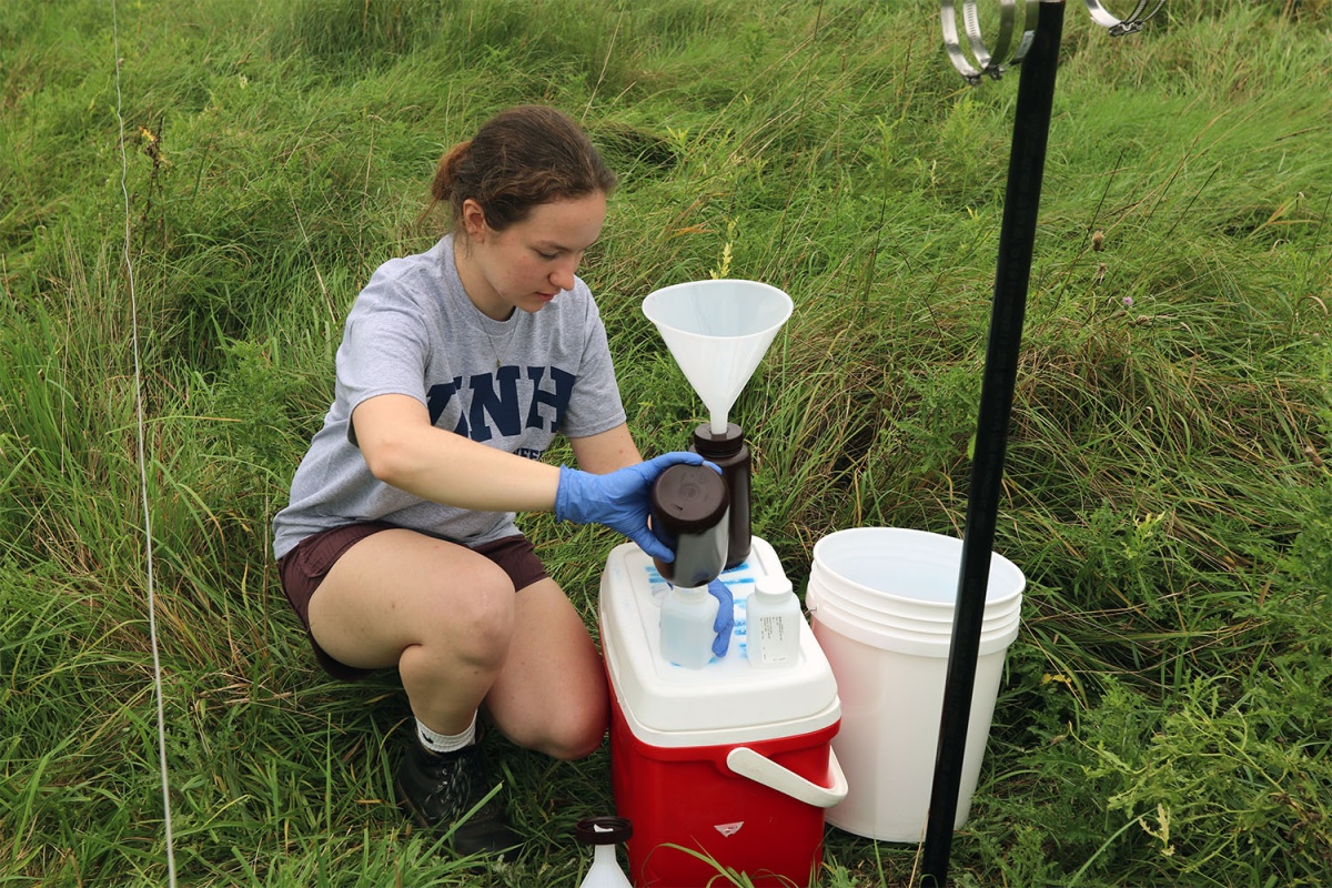 A photo of a woman wearing a CEPS engineering shirt kneeling in a field and gathering samples from a rainwater collection site.
