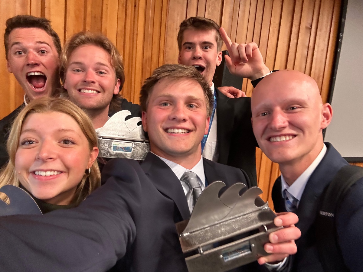A selfie of six UNH students in suits holding wave-shaped trophies