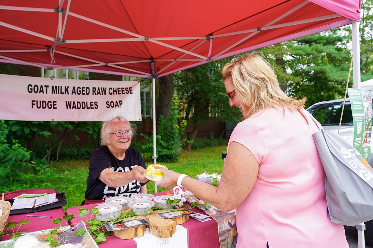 A photo of two women at a farmers market. One woman hands the other a container of goat cheese she is selling. The other woman looks at it.
