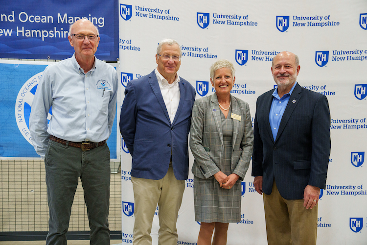 Four people stand in front of a UNH banner