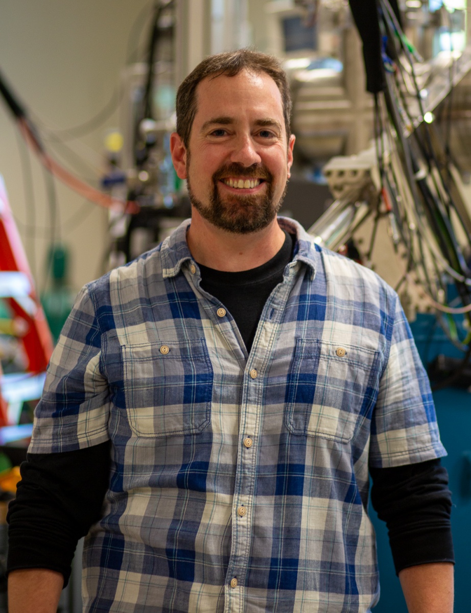 Portrait of white man with beard in front of large scientific equipment