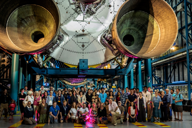 large group of scientists and students pose beneath a rocket ship