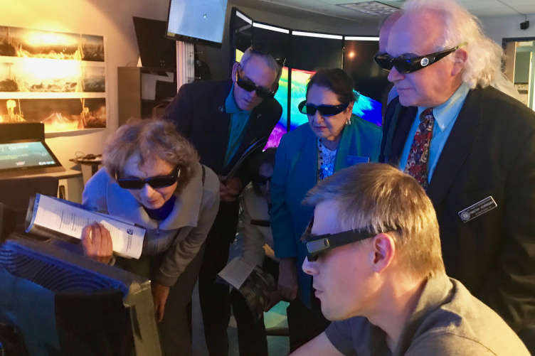 Small group of peope wearing 3D glasses crowd around a computer