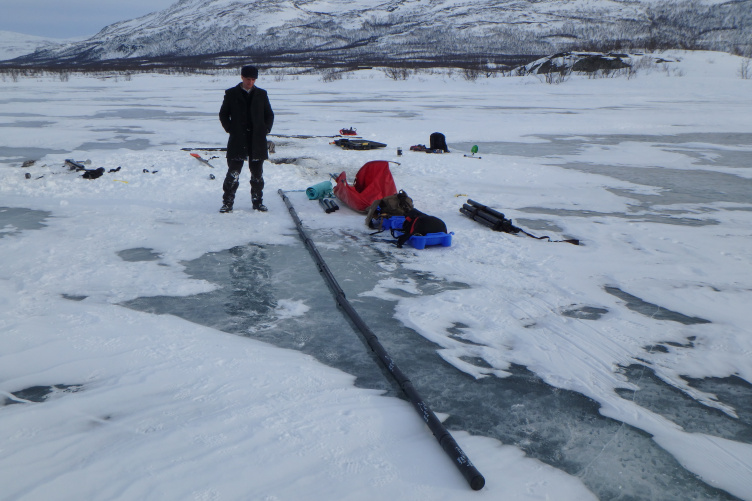 Young man dressed in warm winter clothing stands on frozen lake next to long series of black sediment cores. 