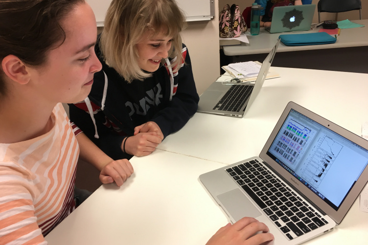 Two young women sit at a table in front of a laptop and smile as they look at colorful graphs on the screen.