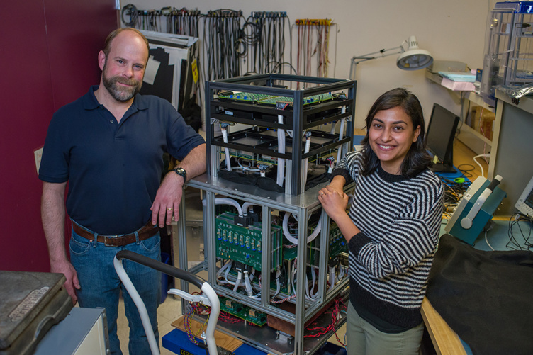 An older man with a beard and a younger woman with dark chin-length hair stand beside a tall cube machine they built.