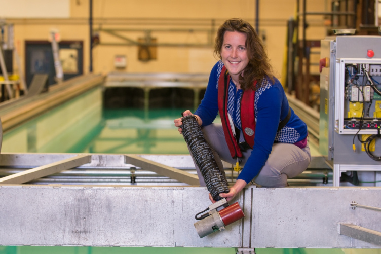 Woman with long brown hair, blue shirt and red life vest stands next to a tank of water while holding a long black cylinder.