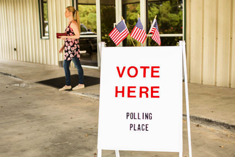 A white sandwich board saying "vote here" is posted outside a building with a woman walking out of the building in the background. A white sandwich board saying "vote here" is posted outside a building with a woman walking out of the building in the background.