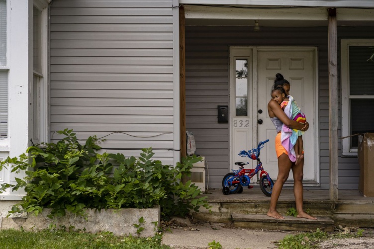 A woman standing in front of her house, holding a child wrapped in a towel.