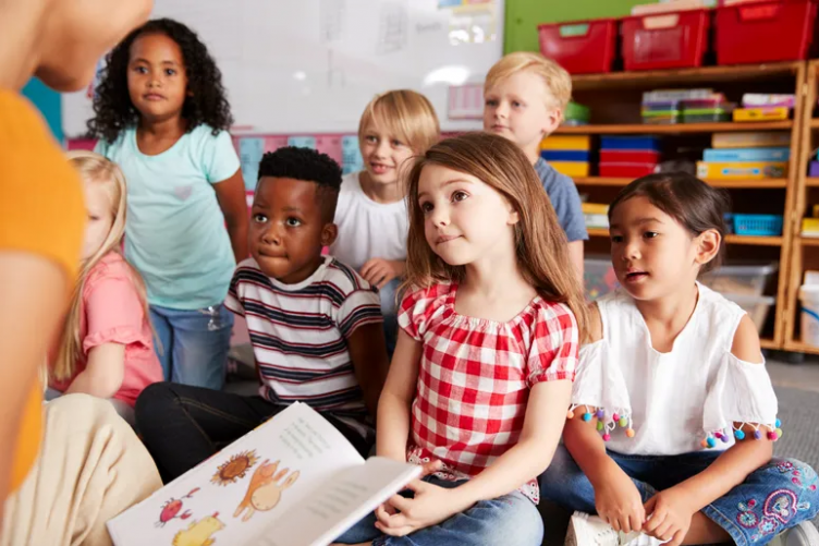 A diverse group of young students listening to their teaching read a book.