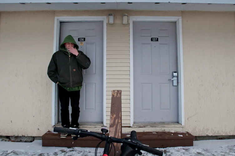 A man stands in the doorway of his apartment smoking a cigarette