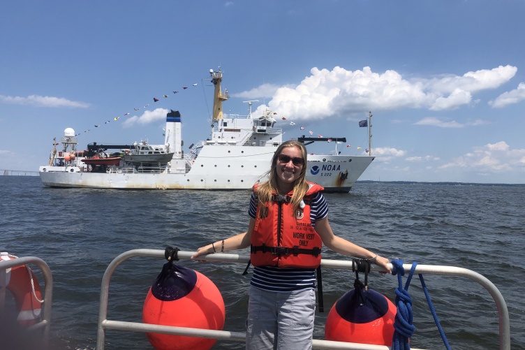 Natalie Cook stands on a ship in front of the Thomas Jefferson hydrographic vessel.