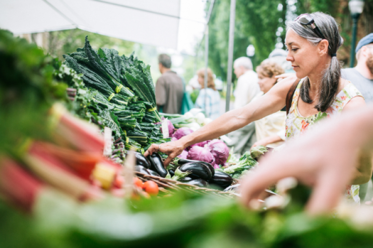 A middle-aged white woman shops at a farmers' market, picking produce from a stall