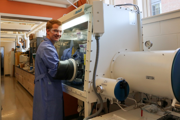 Graduate student wearing protective clothing works with his hands in a large piece of scientific equipment.