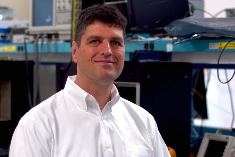 Nathan Schwadron sits in a room full of computer equipment and smiles at the camera.