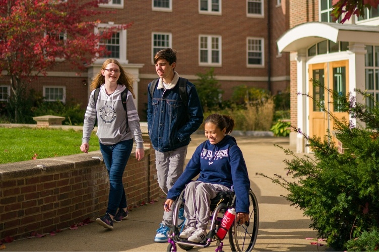 Three UNH students, one in a wheelchair and two standing alongside her, walk and wheel in the courtyard at UNH's Murkland Hall.